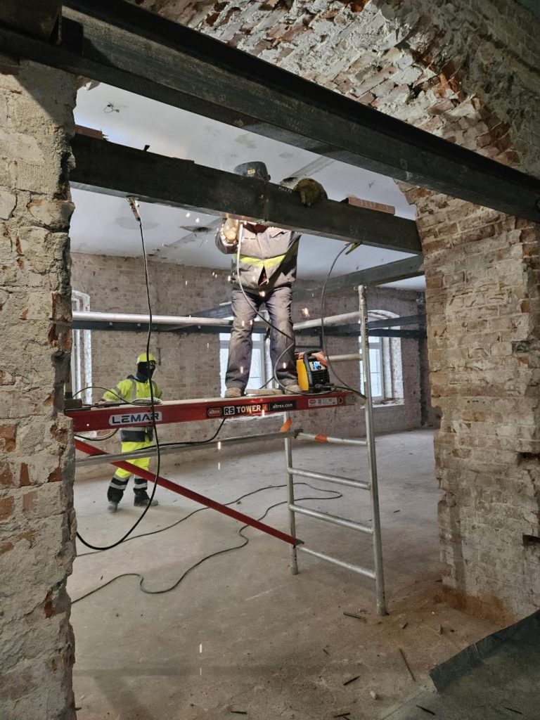 Wnętrze budynku NAC w trakcie przebudowy. Robotnicy montują poziome, metalowe wsporniki wzmacniające konstrukcję przed dalszymi pracami.